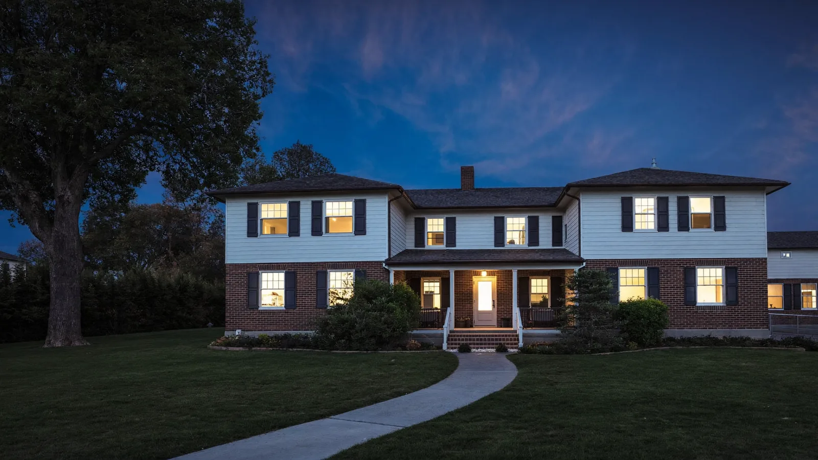 Two-storey brick and siding family home on a wooded lot at blue hour, warm light from the porch and front windows