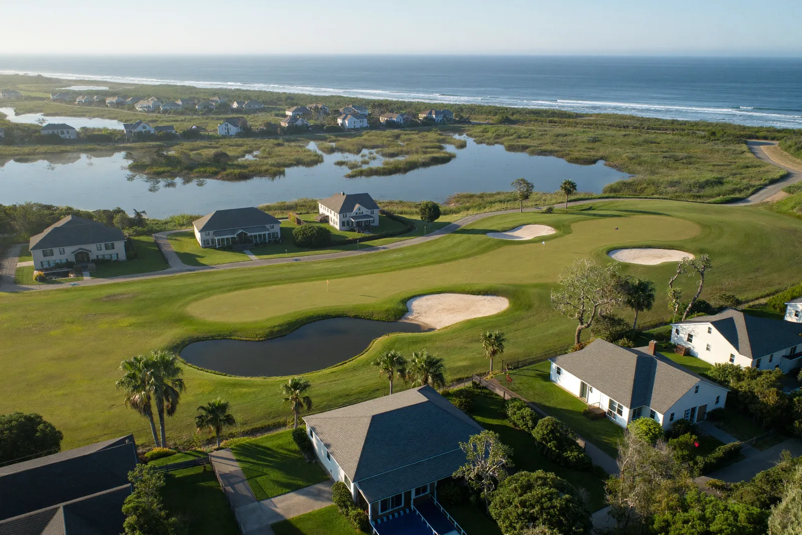 Aerial view of a coastal Grand Strand neighborhood, golf course fairways and bunkers winding between single-family homes with tidal marsh and the Atlantic beyond